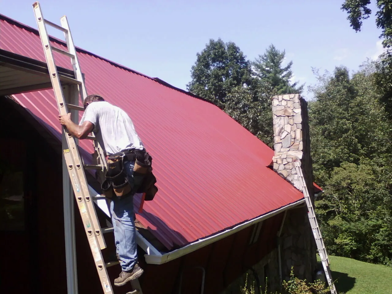 Roofer on a ladder beside a red metal roof and stone chimney — Soffit & Fascia Repair by Roofing Brothers in Lavonia